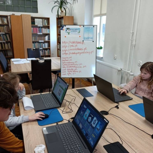 Dzieci siedzą przy laptopach podczas zajęć w bibliotece.